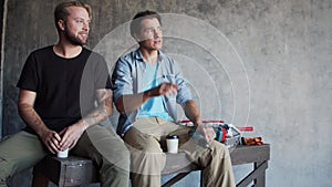 Two friends are resting and talking during a break from interior renovation. Male workers sit on scaffolding and drink