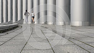 Two friends in sportswear sprint together next to huge pillars, urban workout