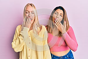 Two friends standing together over pink background bored yawning tired covering mouth with hand