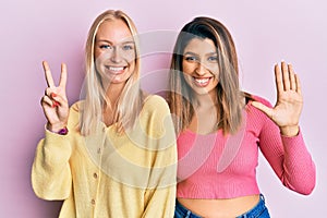 Two friends standing together over pink background showing and pointing up with fingers number seven while smiling confident and