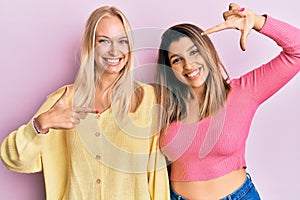 Two friends standing together over pink background smiling making frame with hands and fingers with happy face