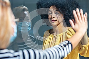 Two friends touching her hands separated by a glass window for social distancing during the corona virus pandemic