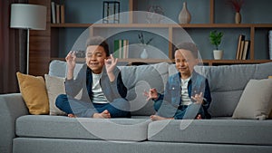 Two funny little African American ethnic children boys brothers siblings kids meditating together sitting on couch in