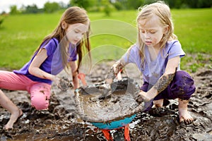 Two funny little girls playing in a large wet mud puddle on sunny summer day. Children getting dirty while digging in muddy soil.