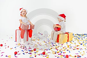 Two funny little kids in Santa hat sitting on gift boxes. Isolated on white background, confetti on a floor. Christmas and new