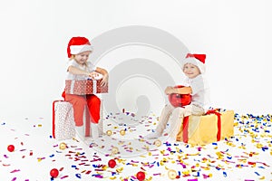 Two funny little kids in Santa hat sitting on gift boxes. Isolated on white background, confetti on a floor. Christmas and new