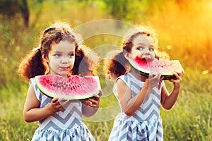 Two funny little sisters eating watermelon outdoors on warm and sunny summer day. Healthy organic food for little kids. Twins girl