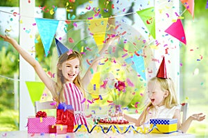 Two funny little sisters having birthday party at home, blowing candles on birthday cake