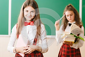 Two funny schoolgirls in school uniform are standing with books on the background of the school board