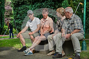 Two generation of peoples are sitting on a park bench at a minigolf court. They are waiting that they can use the court..