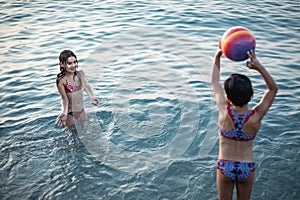 two gils in swimsuits standing in the water and playing with colourful ball in the sea