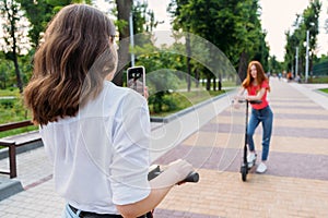 Two girl friends riding push e scooter and do selfie via their smartphones outdoors. Group gen z young people using