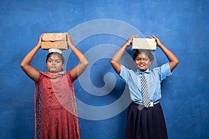 Two girl kids with books and bricks on each other head looking at camera - concept poverty and conflict or necessary