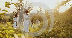 Two girl teenagers in white dresses and straw hats with flowers bouquet walking on nature at sunny summer day