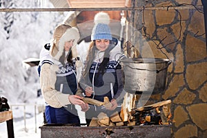 Two girlfriends cooking in the winte nature on a fire in the boiler. Girls bask in the fire in winter.