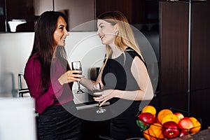 Two girlfriends in elegant clothes relaxing after shopping, drinking wine, laughing and gossiping in the kitchen