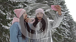 Two girlfriends with a good mood make selfie using a smartphone while standing in the winter forest. slow motion