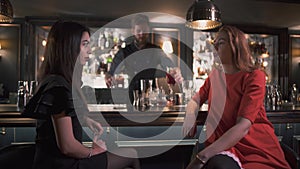 Two girlfriends sitting near bar counter having interesting conversation close up. Bartender gives cocktails to them