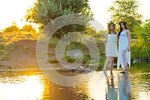 Two girlfriends, young girls in white dresses and flower wreaths on their heads, standing in a river of water, near the shore,