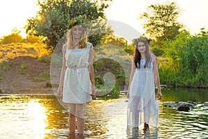 Two girlfriends, young girls in white dresses and flower wreaths on their heads, standing in a river of water, near the shore,