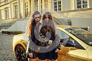 Two girls on a background of a yellow car. Beautiful brunettes near the cottage arrived on an expensive car. Famous girls.