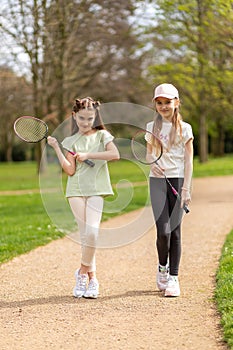 Two girls with badminton rackets on a park path.