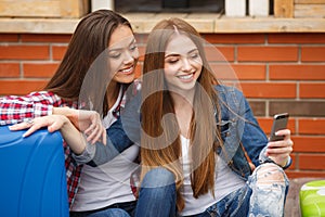 Two girls with bags reading text message while sitting at the station