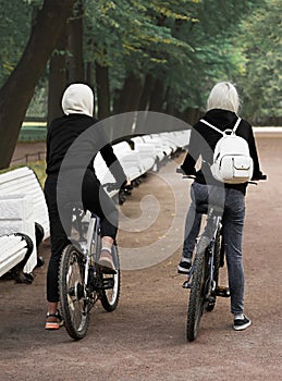 Two girls on bicycles on a walk in a deserted park. Alley, benches, path sprinkled with gravel. Photo.
