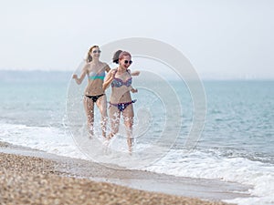 Two girls in bikini running along seashore splashing sea water