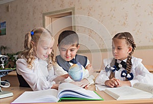 Two girls and a boy at school at a Desk studying geography watching the globe. The concept of education in the school