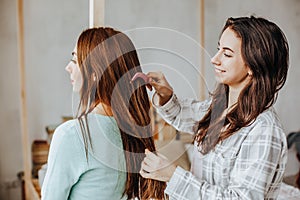 Two girls braid their hair at the window. Woman makes a braid to her friend. Hair weaving hairstyles. Girlfriend braids her hands