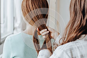 Two girls braid their hair at the window. Woman makes a braid to her friend. Hair weaving hairstyles. Girlfriend braids her hands