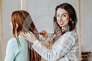 Two girls braid their hair at the window. Woman makes a braid to her friend. Hair weaving hairstyles. Girlfriend braids her hands