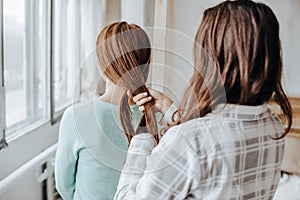 Two girls braid their hair at the window. Woman makes a braid to her friend. Hair weaving hairstyles. Girlfriend braids her hands