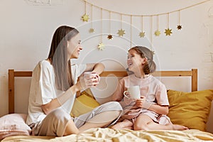 Two girls, cheerful older and younger sister drinking tea and talking while spending time together in bedroom