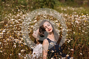 Two girls with closed eyes in dark blue and white dresses in sunny day sitting in chamomile field