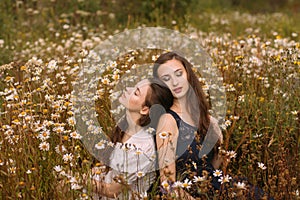 Two girls with closed eyes in dark blue and white dresses in sunny day sitting in chamomile field