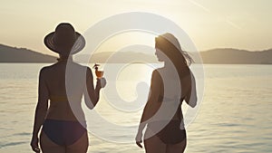 Two girls with cocktails in their hands walking on the beach by the ocean.