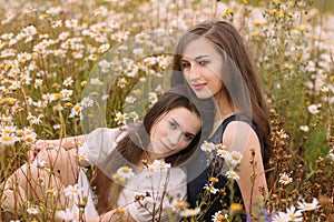 Two girls in dark blue and white dresses in sunny day sitting in chamomile field