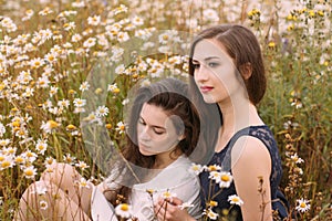 Two girls in dark blue and white dresses in sunny day sitting in chamomile field