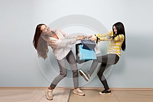 Two girls dressed in nice casual clothes pull and take each other shopping bags on the white background