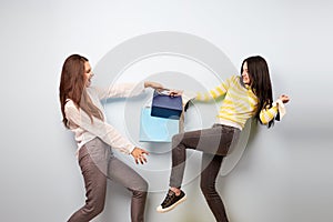 Two girls dressed in nice stylish clothes are pulling each other bags after shopping on the white background