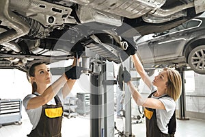 Two girls fixing lifted auto undercarriage, using wrenches.