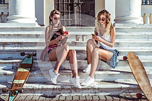 Two girls girlfriends in summer rest on steps. Nearby are skateboards, longboards. In hands of holding smartphones
