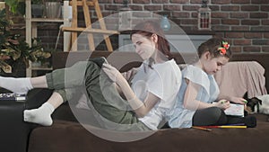 Two girls inside are sitting on cozy sofa back to back and cheerfully drawing