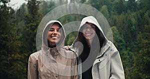 Two girls in light jackets in hoods rejoice in the coming rain and look at the camera in a mountain forest