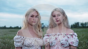 Two girls in light summer dresses stand in a field and look into the camera