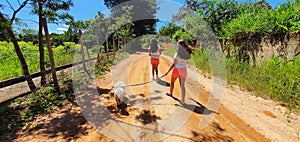 Two girls with long hair, pink shorts and a white shirt, walk with a white poodle named Nina, along the dirt road, flanked by