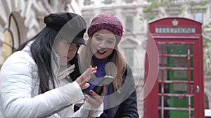 Two girls look at the mobile phone on a London street with a red telephone booth