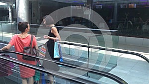 Two girls moving down the stairs on escalator in shopping mall, friends are shopping in a store, women making purchases